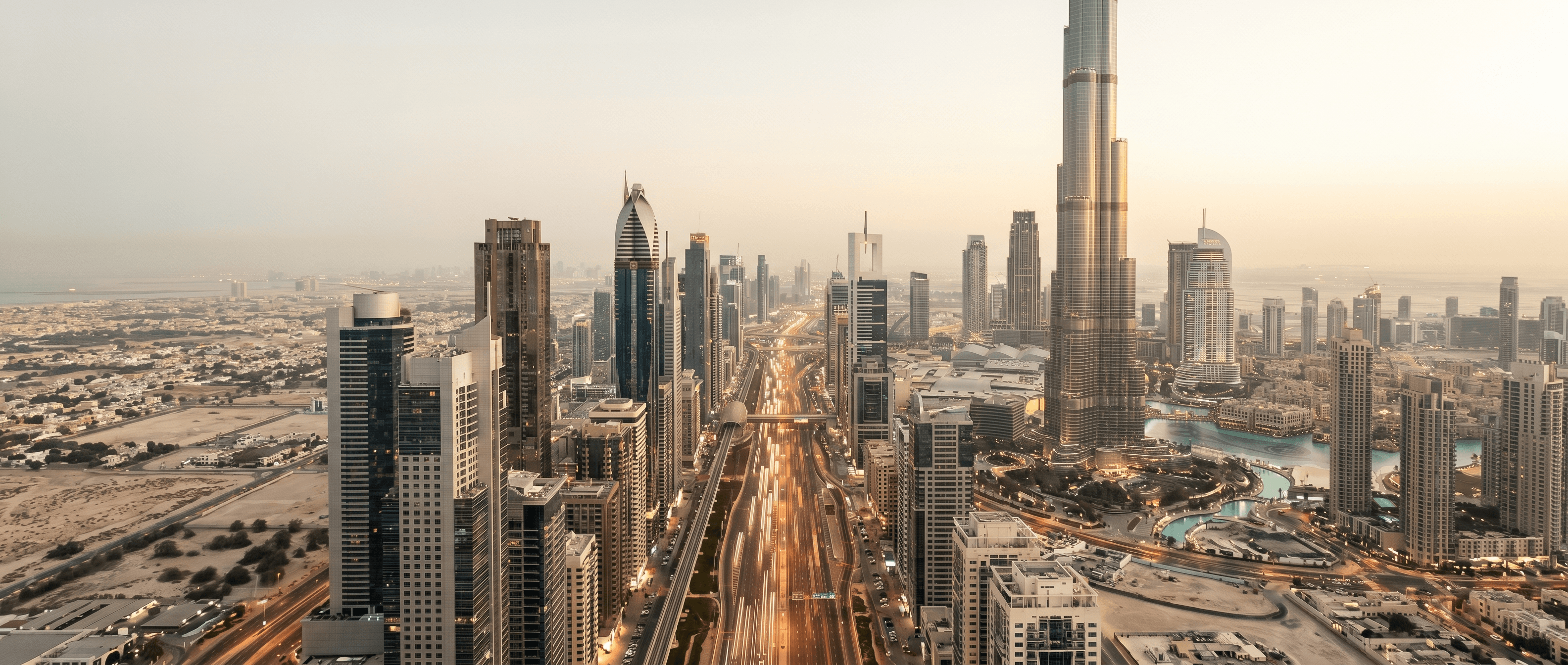 An aerial panoramic of Sheikh Zayed Road at golden hour — traffic streams beneath the towers, the Burj Khalifa visible to the right.