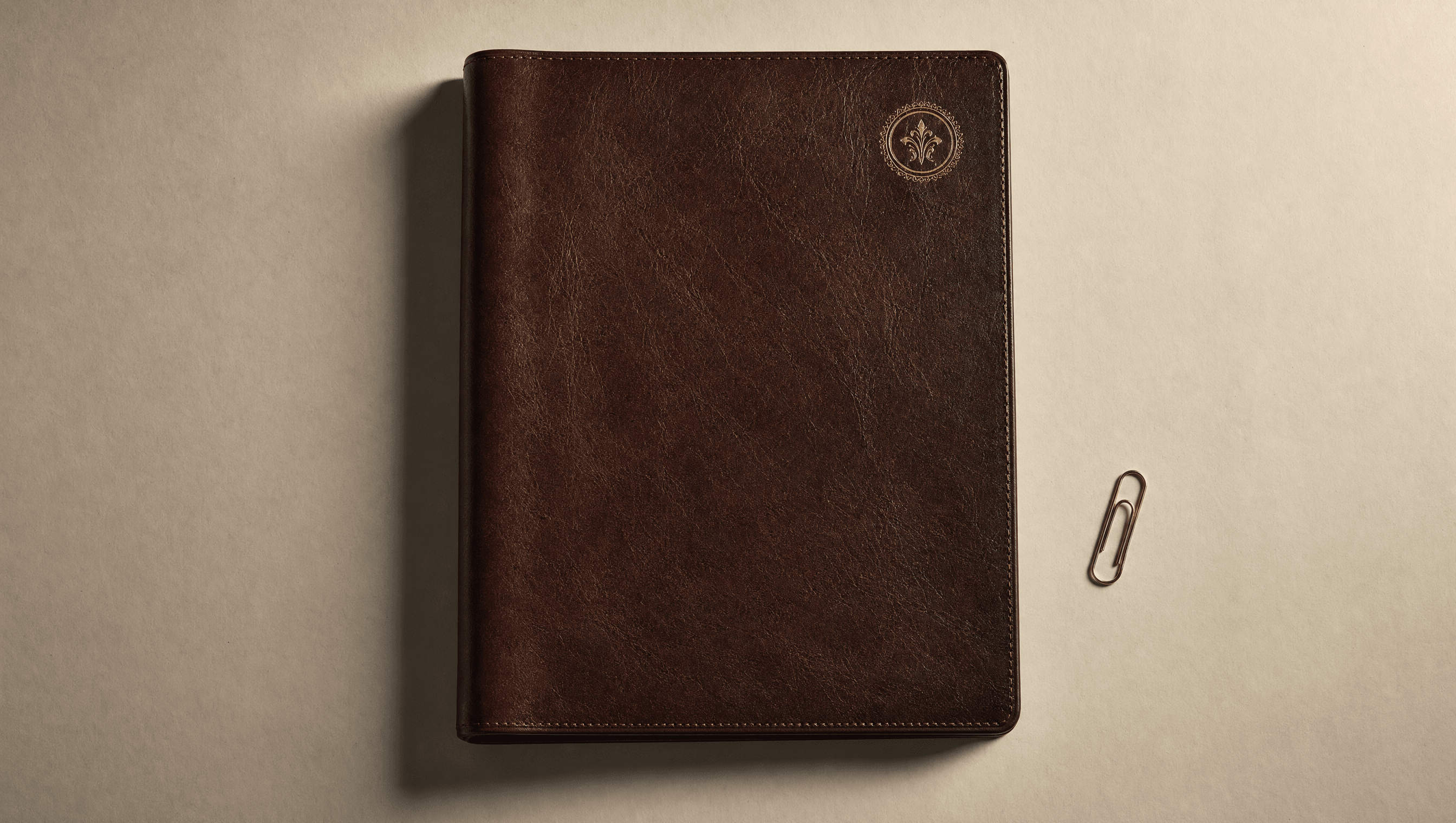 A closed leather folder embossed with a small gold-foil monogram, lying on cream paper with a brass paperclip beside it.